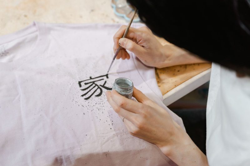 Hands painting a black character onto a pale tee with a fine brush and ink