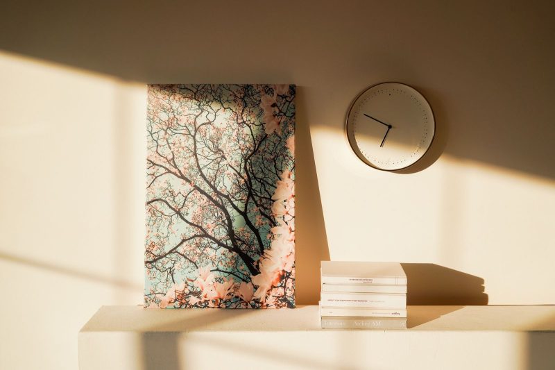 Canvas print of pink blossoms leaning on a sunlit shelf beside a wall clock