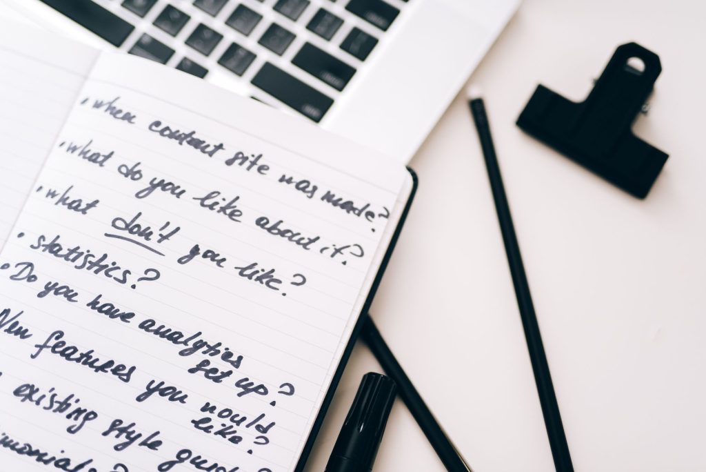 An open notebook with a handwritten list of questions beside a laptop