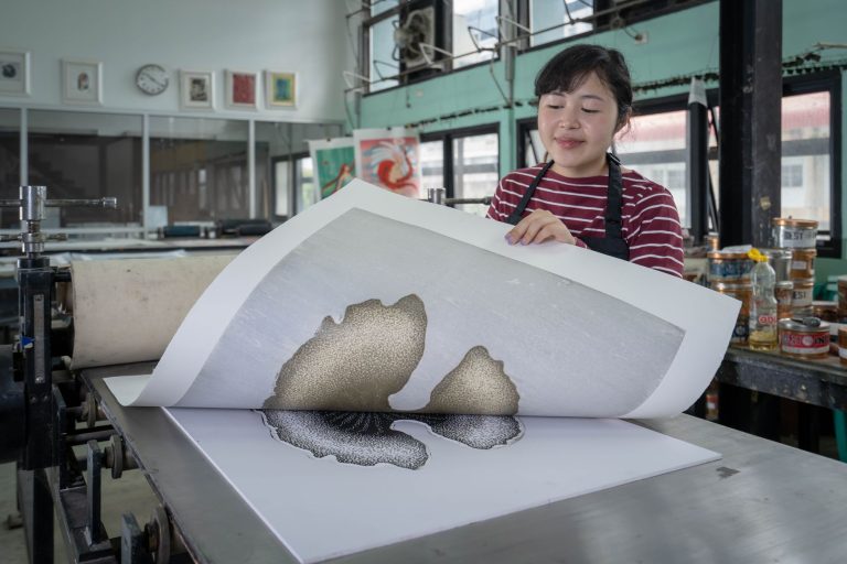 An artist examining a numbered print fresh off the press in a printmaking studio