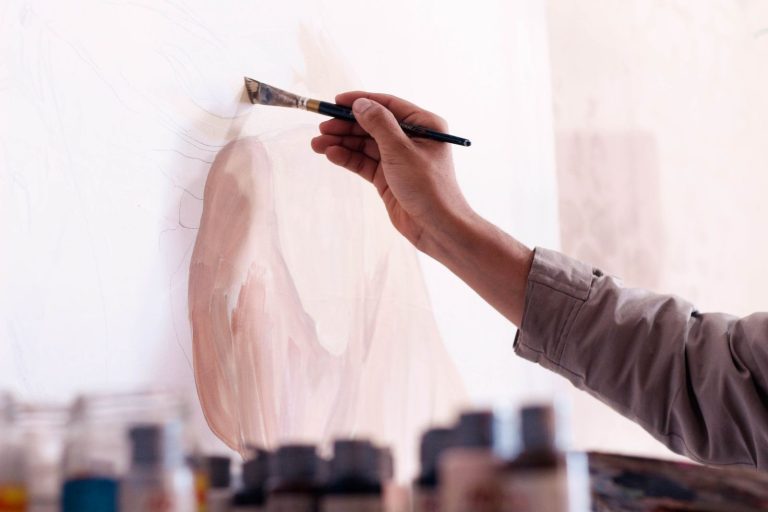 Artist's hand painting soft pink shapes on a large canvas with paint jars below